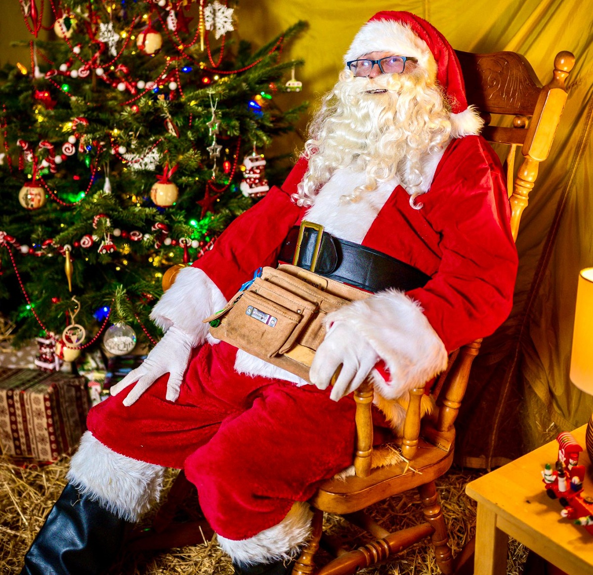 Santa Claus dressed in a traditional red suit with white fur trim, sitting in a wooden rocking chair beside a decorated Christmas tree. Santa has a large black belt and a tan utility pouch around the waist, with wrapped presents visible under the tree and festive decorations in the background.