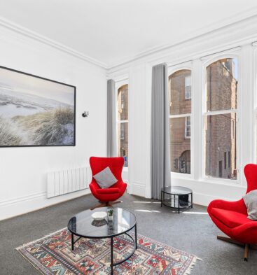 A living room featuring a large framed beach artwork, two vibrant red modern armchairs, a glass coffee table over an area rug, and three tall windows.