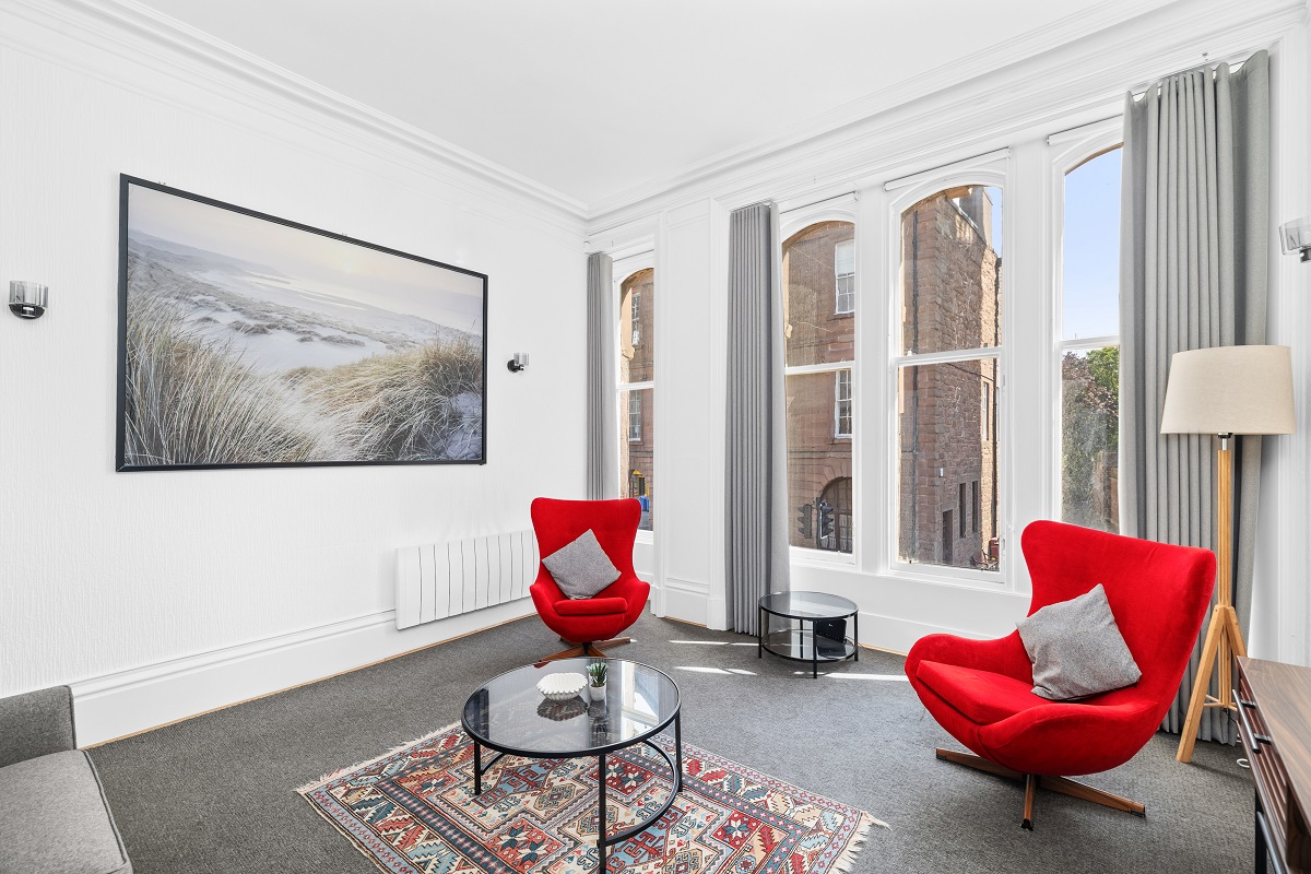 A living room featuring a large framed beach artwork, two vibrant red modern armchairs, a glass coffee table over an area rug, and three tall windows.