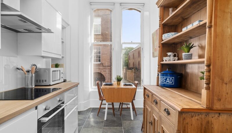 A bright, narrow kitchen with modern white cabinetry, a wood counter, a small wooden dining table, and a large rustic wooden hutch on the right.