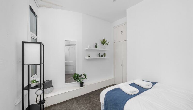 A minimalist white bedroom with a double bed, dark gray carpet, a black shelving unit, and floating white shelves with small plants.
