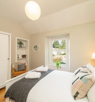A neutral-toned bedroom with a white bed and headboard, a white wardrobe, a small sitting area with a wicker chair, and a window overlooking greenery.