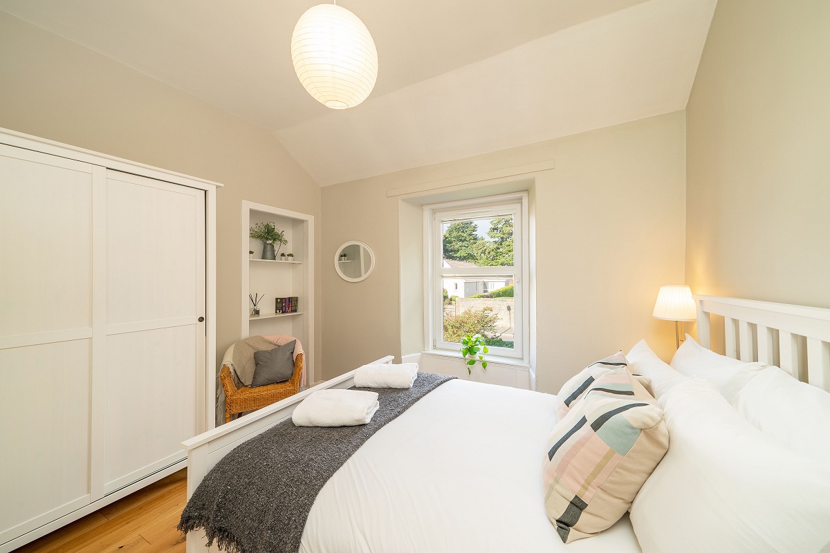 A neutral-toned bedroom with a white bed and headboard, a white wardrobe, a small sitting area with a wicker chair, and a window overlooking greenery.