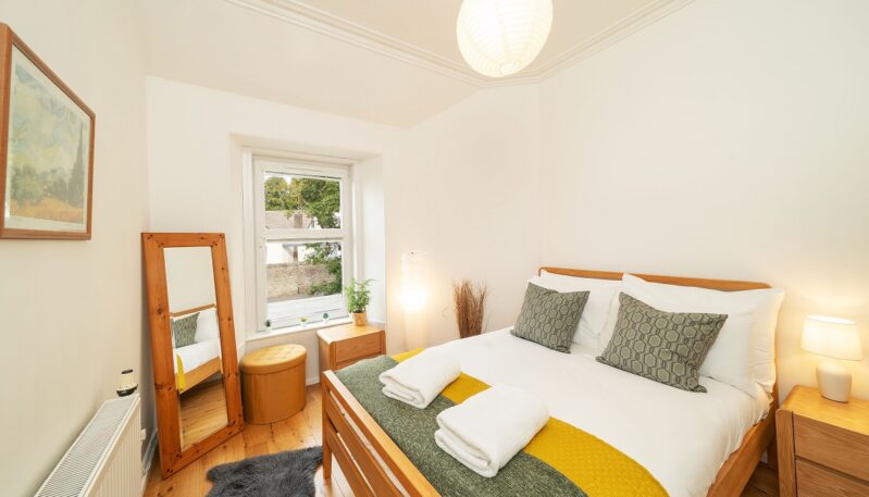 A bright bedroom with white walls, a wooden bed frame, a yellow and green throw blanket, a full-length wooden mirror, and a small window.