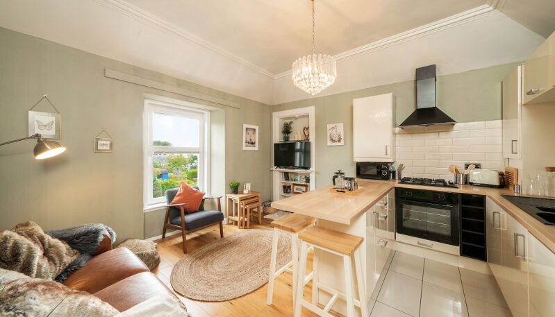 A compact open-plan living room and kitchen featuring pale green walls, a leather couch, a small armchair, and a kitchen island with white cabinetry and wooden counters.