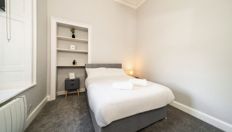 A minimalist bedroom featuring a gray fabric double bed, a matching gray nightstand, a built-in white shelf niche, and dark gray carpet.