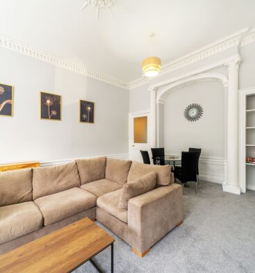 A spacious living and dining room with a large tan sectional sofa, a wooden coffee table, ornate white crown molding, and a small dining area visible through an arched doorway.
