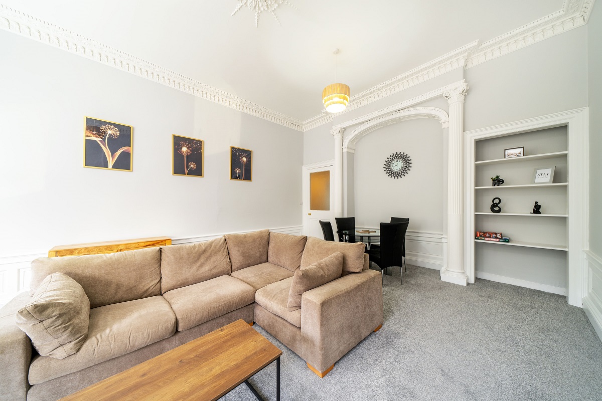 A spacious living and dining room with a large tan sectional sofa, a wooden coffee table, ornate white crown molding, and a small dining area visible through an arched doorway.
