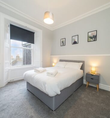 A bright, modern bedroom with pale gray walls, a gray fabric bed, a large bay window with a black roller blind, and simple framed artwork above the bed.