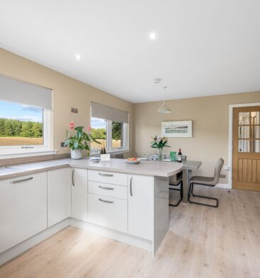 A bright, modern kitchen and dining area with white glossy cabinets, wood-look laminate flooring, and a large window offering a view of a field and forest.