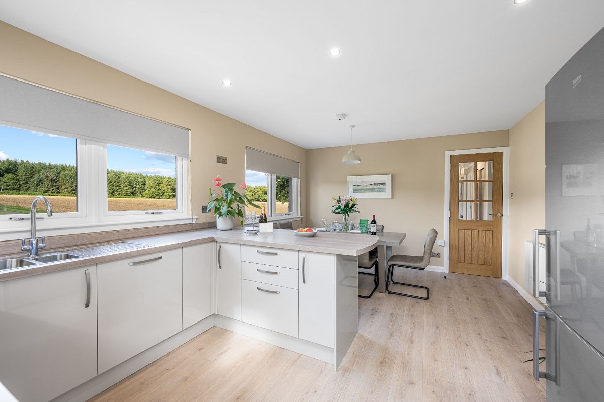 A bright, modern kitchen and dining area with white glossy cabinets, wood-look laminate flooring, and a large window offering a view of a field and forest.
