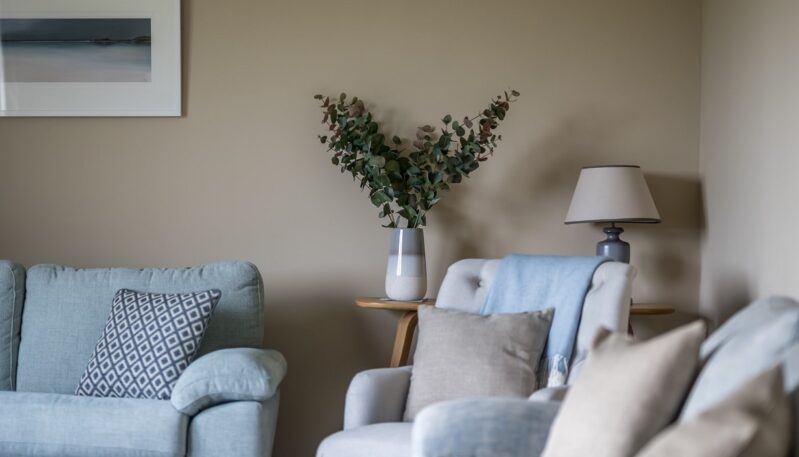 A close-up of a cozy living room with pale taupe walls, soft blue and light gray upholstered seating, and a vase of eucalyptus on a side table next to a lamp.