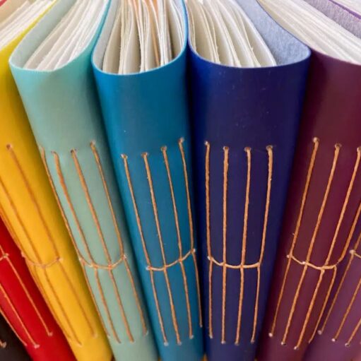 A close-up, angled view of the spines of several handmade journals lined up, showing a gradient of bright colors (red, yellow, turquoise, blue, purple, and pink) with exposed, natural-toned coptic binding stitches.