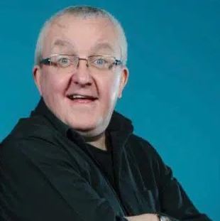A headshot of a smiling man with glasses and short, white hair wearing a black shirt, looking slightly off-center against a bright blue background.