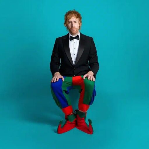 A full-length photo of a man with red hair wearing a black tuxedo and bow tie on top, paired with brightly colored clown or elf pants (red, green, and blue) and red elf shoes, sitting down against a solid teal background.