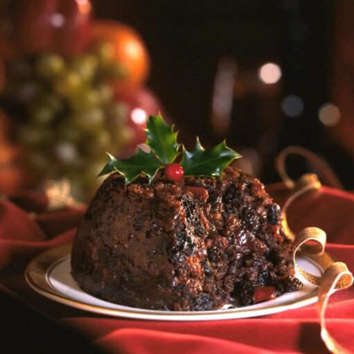 A traditional, rich, dark Christmas pudding served on a white plate with gold trim, garnished with a sprig of holly and a red berry, resting on red fabric with a gold ribbon, and blurred fruit in the background.