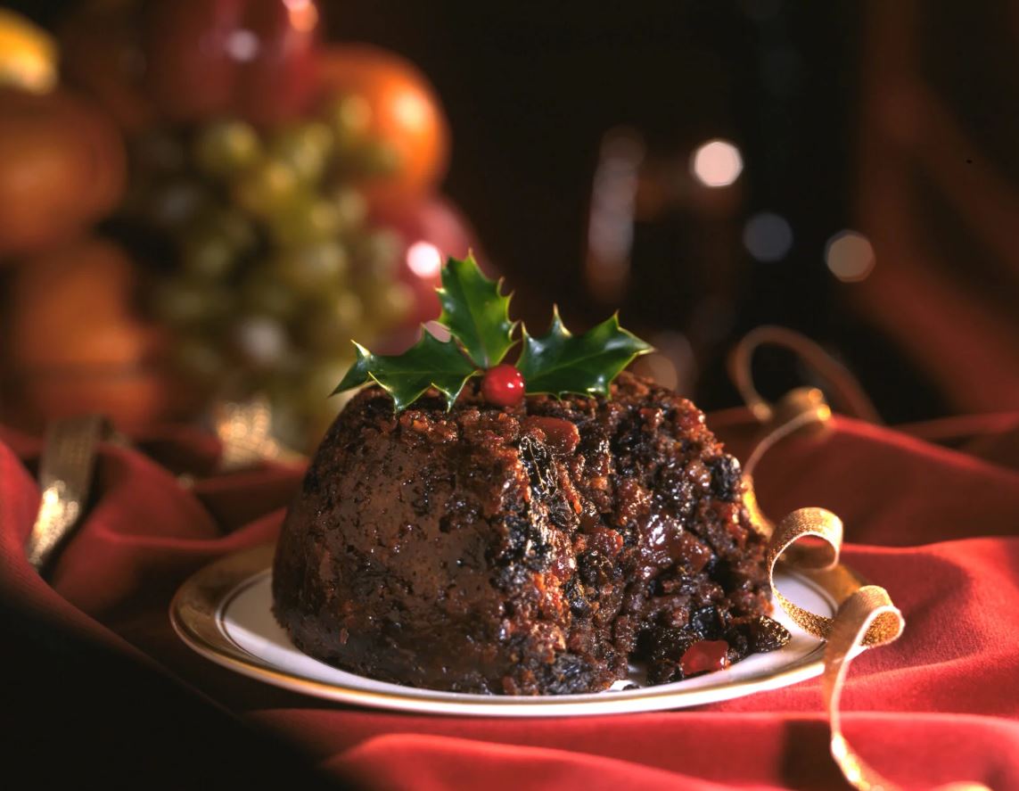 A traditional, rich, dark Christmas pudding served on a white plate with gold trim, garnished with a sprig of holly and a red berry, resting on red fabric with a gold ribbon, and blurred fruit in the background.