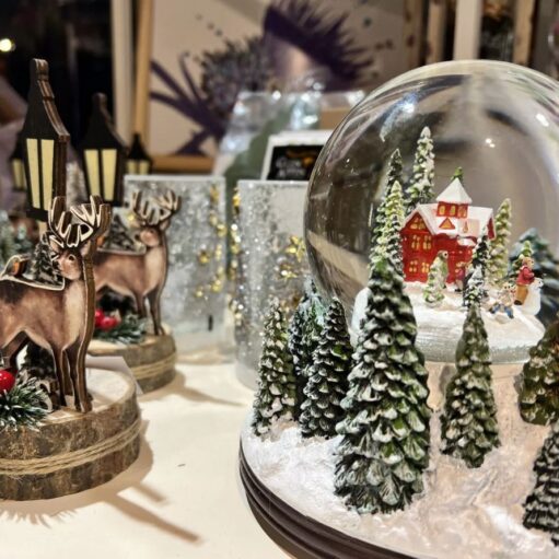 A close-up display of Christmas decorations including a large snow globe featuring a miniature red house and snow-covered evergreen trees, alongside wooden reindeer figures and glittery silver candle holders.