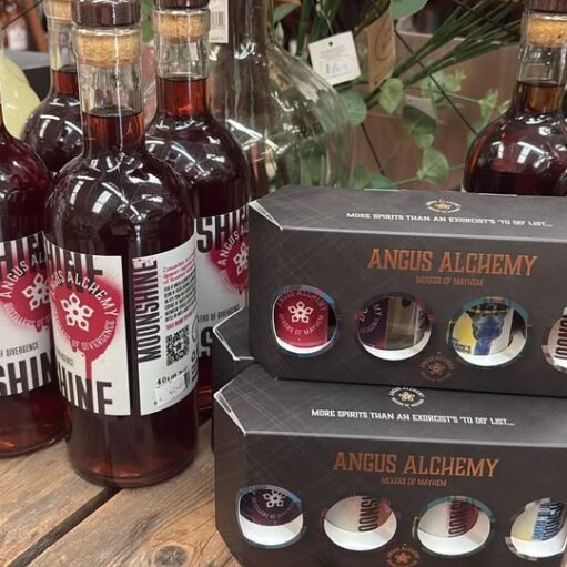 A display of bottles of Angus Alchemy Moonshine (a dark red spirit) and black cardboard gift sets labeled "Mixes of Mayhem" resting on a rustic wooden barrel or table in a shop.