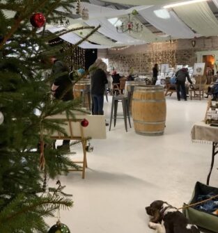 An interior view of the Rottal Market, possibly a Christmas fair, with a decorated Christmas tree in the foreground. People are mingling around high tables made from wooden barrels, and various vendor stalls are visible in the background against stone and white draped walls. A dog is lying down near the right side of the image.