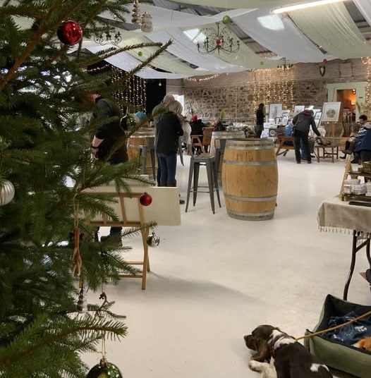 An interior view of the Rottal Market, possibly a Christmas fair, with a decorated Christmas tree in the foreground. People are mingling around high tables made from wooden barrels, and various vendor stalls are visible in the background against stone and white draped walls. A dog is lying down near the right side of the image.