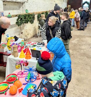 A lively indoor market scene at Pathhead, featuring a vendor stall with colorful novelty items, toys, and Christmas decorations. A woman is speaking to a man on the left, while two children in winter hats and jackets look at the items on the table. Other shoppers are visible in the background, and the walls are decorated with Christmas garlands.