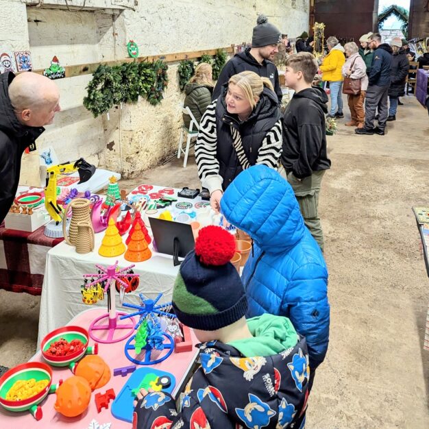 A lively indoor market scene at Pathhead, featuring a vendor stall with colorful novelty items, toys, and Christmas decorations. A woman is speaking to a man on the left, while two children in winter hats and jackets look at the items on the table. Other shoppers are visible in the background, and the walls are decorated with Christmas garlands.