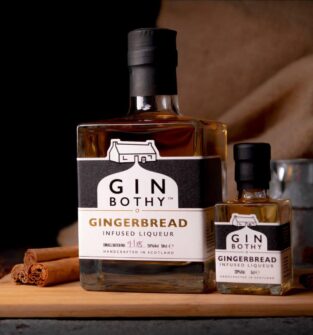 A professional studio photograph featuring two bottles of Gin Bothy Gingerbread Infused Liqueur, a full-sized bottle and a miniature, resting on a wooden board alongside cinnamon sticks.