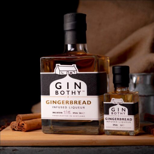A professional studio photograph featuring two bottles of Gin Bothy Gingerbread Infused Liqueur, a full-sized bottle and a miniature, resting on a wooden board alongside cinnamon sticks.