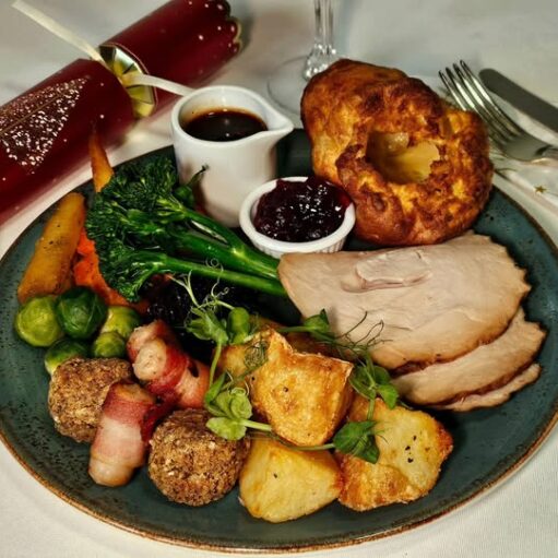 Traditional festive roast dinner served on a plate with sliced turkey, roast potatoes, Yorkshire pudding, stuffing, pigs in blankets, seasonal vegetables, cranberry sauce and gravy, with Christmas crackers on the table.