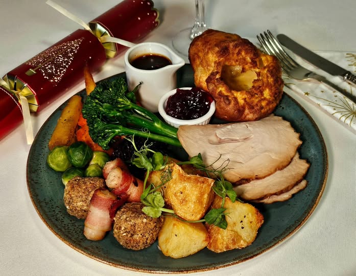 Traditional festive roast dinner served on a plate with sliced turkey, roast potatoes, Yorkshire pudding, stuffing, pigs in blankets, seasonal vegetables, cranberry sauce and gravy, with Christmas crackers on the table.