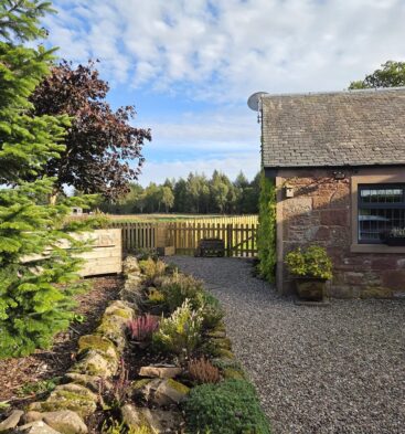 The exterior of a traditional stone building with a slate roof, a gravel path, a rock garden, and a wooden fence leading to a wooded area under a blue sky.