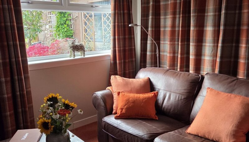 A cosy living area featuring a brown leather sofa with orange pillows, a dark coffee table with a vase of flowers, and windows framed by orange and brown tartan curtains.