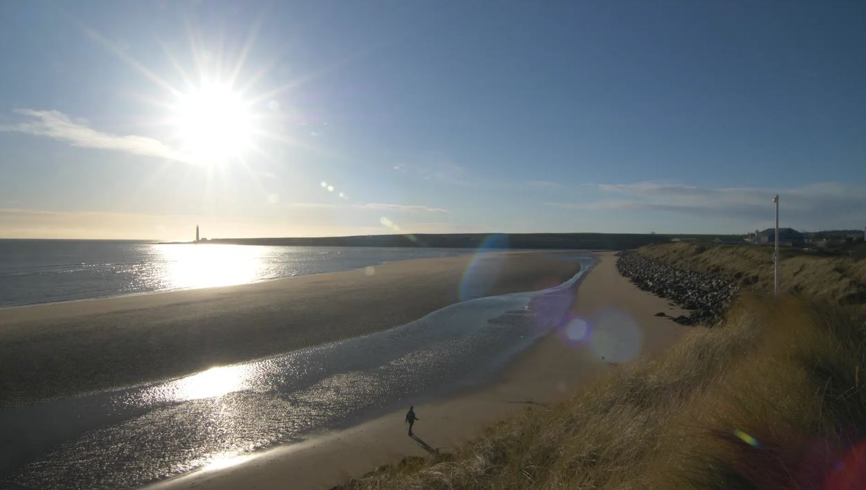 A wide-angle landscape photo of a sunny beach scene with a bright sunburst in the upper left. A small figure walks along the wet sand where a stream meets the sea. There are tall grassy dunes on the right and a distant lighthouse on the horizon over the water.
