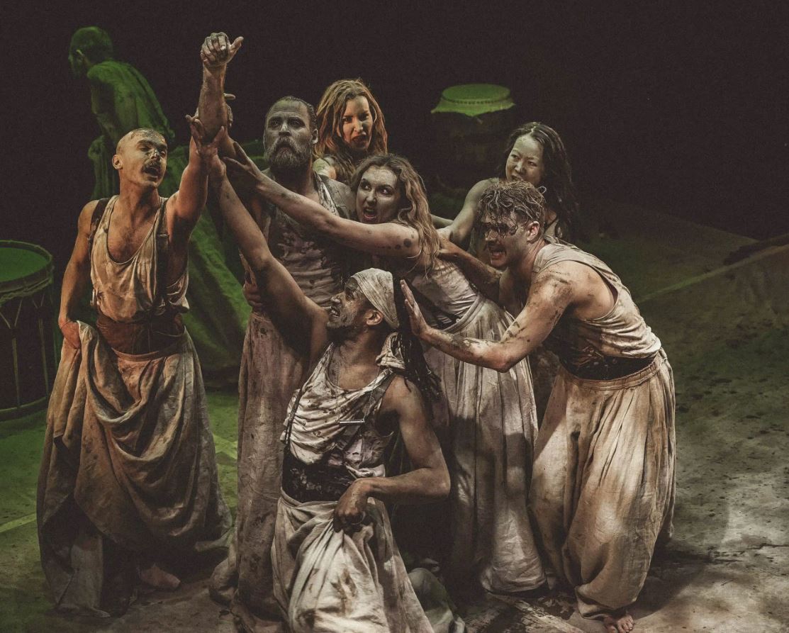 A dramatic theatrical scene featuring seven performers, all covered in dark grime or mud and wearing tattered, dirty white tank tops and robes. They are grouped closely, looking up and reaching their arms skyward in an intense emotional tableau. The lighting is dim, emphasizing the performers and their expressions.
