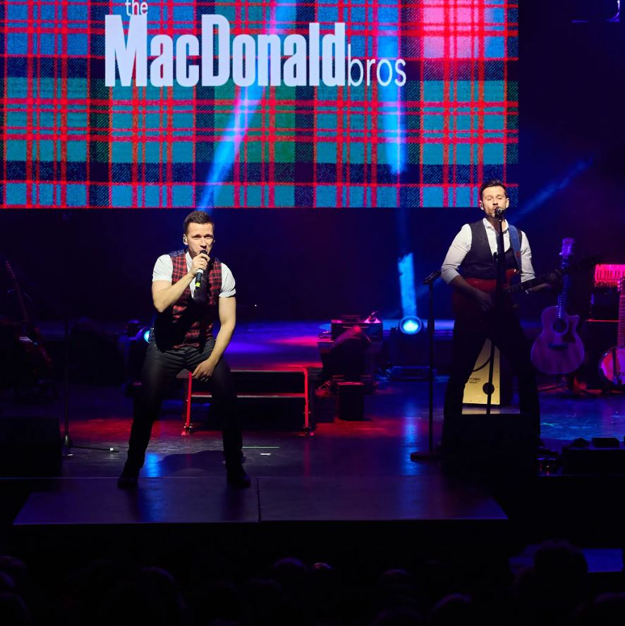Two male musicians, the MacDonald Bros, perform on a stage. One is singing into a microphone stand at the front, and the other is singing while playing a guitar further back. They are wearing dark vests over white shirts. Behind them is a large screen displaying the words "the MacDonald bros" over a red, blue, and green tartan pattern.