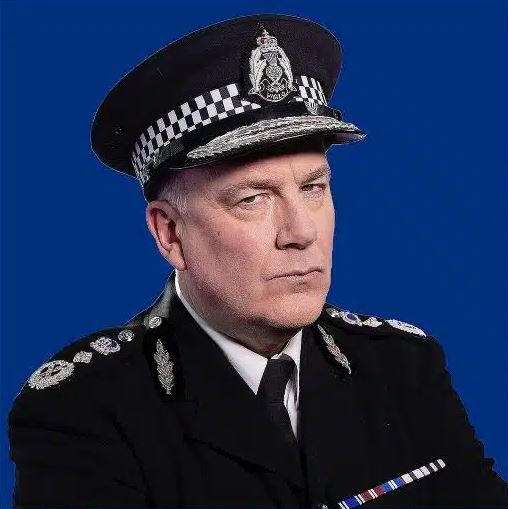 A headshot of a stern-looking man wearing a formal police or military uniform, including a peaked cap with insignia and a checkered band, and a dark jacket with silver epaulets and a ribbon bar. The background is a solid blue.