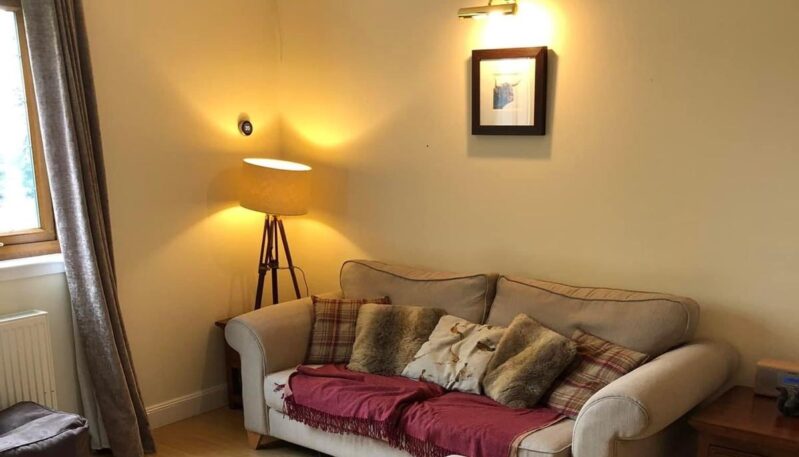 Cosy living area featuring a beige two-seater sofa with tartan and faux-fur cushions, a red throw, and a tripod floor lamp next to a window.