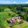 Aerial view of a large, modern lodge building with a brown roof and paved driveway, surrounded by lush green lawns and dense woodland.