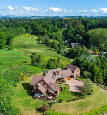 Aerial view of a large, modern lodge building with a brown roof and paved driveway, surrounded by lush green lawns and dense woodland.