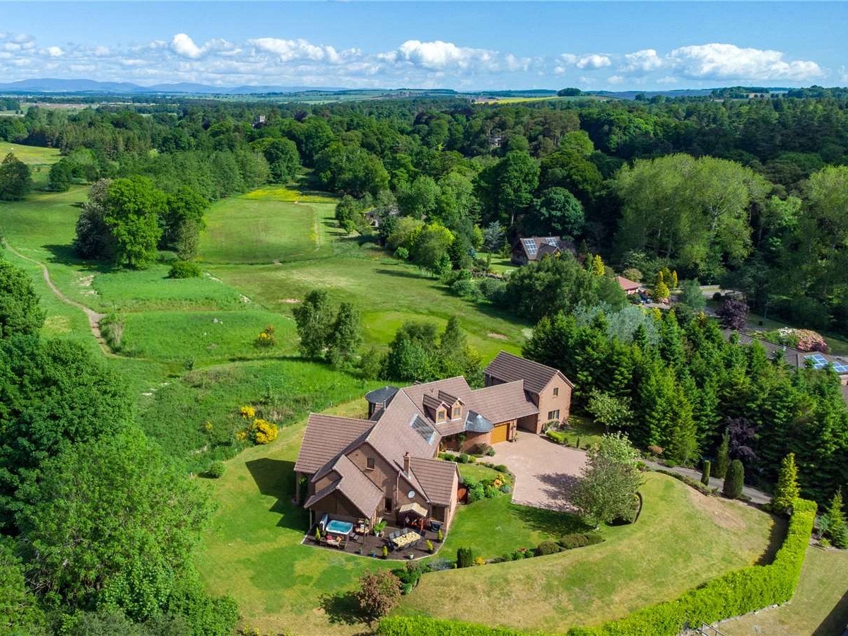 Aerial view of a large, modern lodge building with a brown roof and paved driveway, surrounded by lush green lawns and dense woodland.