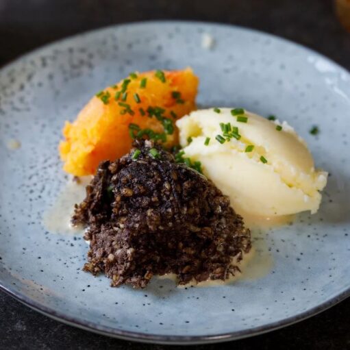 A portion of haggis (dark brown, crumbly texture) served with mashed potatoes (creamy white) and neeps (mashed swede/turnip, orange), all topped with chopped chives and a drizzle of sauce, presented on a light blue-grey speckled plate.