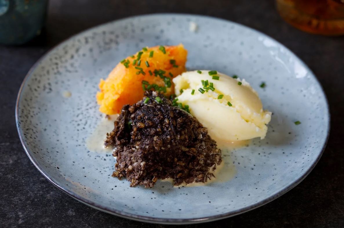 A portion of haggis (dark brown, crumbly texture) served with mashed potatoes (creamy white) and neeps (mashed swede/turnip, orange), all topped with chopped chives and a drizzle of sauce, presented on a light blue-grey speckled plate.
