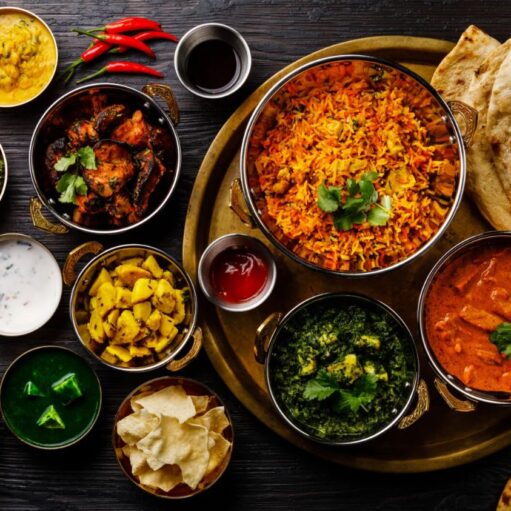 An overhead flat lay of a variety of colorful Indian dishes and accompaniments. A large brass serving platter in the center holds a bowl of biryani and another bowl of curry. Around the platter are several smaller brass bowls containing different curries, rice, sides, and sauces, including chutneys, yogurt, greens, and yellow potatoes, along with papadums, naan bread, and garnishes like lime slices and chili peppers.