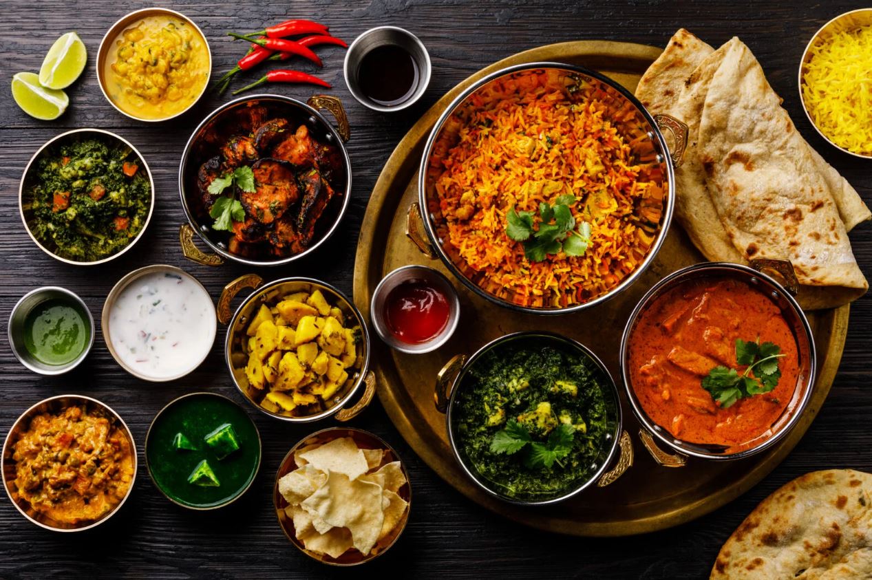 An overhead flat lay of a variety of colorful Indian dishes and accompaniments. A large brass serving platter in the center holds a bowl of biryani and another bowl of curry. Around the platter are several smaller brass bowls containing different curries, rice, sides, and sauces, including chutneys, yogurt, greens, and yellow potatoes, along with papadums, naan bread, and garnishes like lime slices and chili peppers.