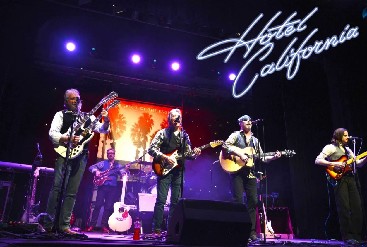 A band on a dimly lit stage performing, with four members visible playing guitars and singing. The background features a large screen displaying an image of palm trees at sunset and text reading "THE SPIRIT OF THE EAGLE." In the upper right corner, "Hotel California" is written in a cursive blue neon-style font.