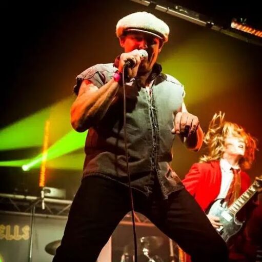 A male singer on stage wearing a flat cap and a sleeveless denim or canvas shirt, leaning into a microphone. Behind him to the right, a guitarist in a red jacket with long hair is partially visible, playing a silver electric guitar. The stage is lit with bright green and red lights.