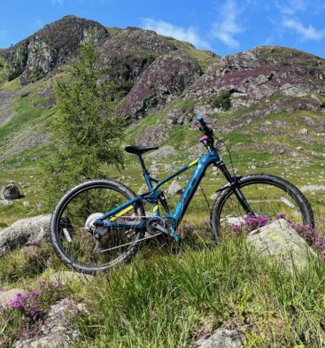 A blue and yellow mountain bike placed among rocks and purple heather in a rugged mountainous landscape.