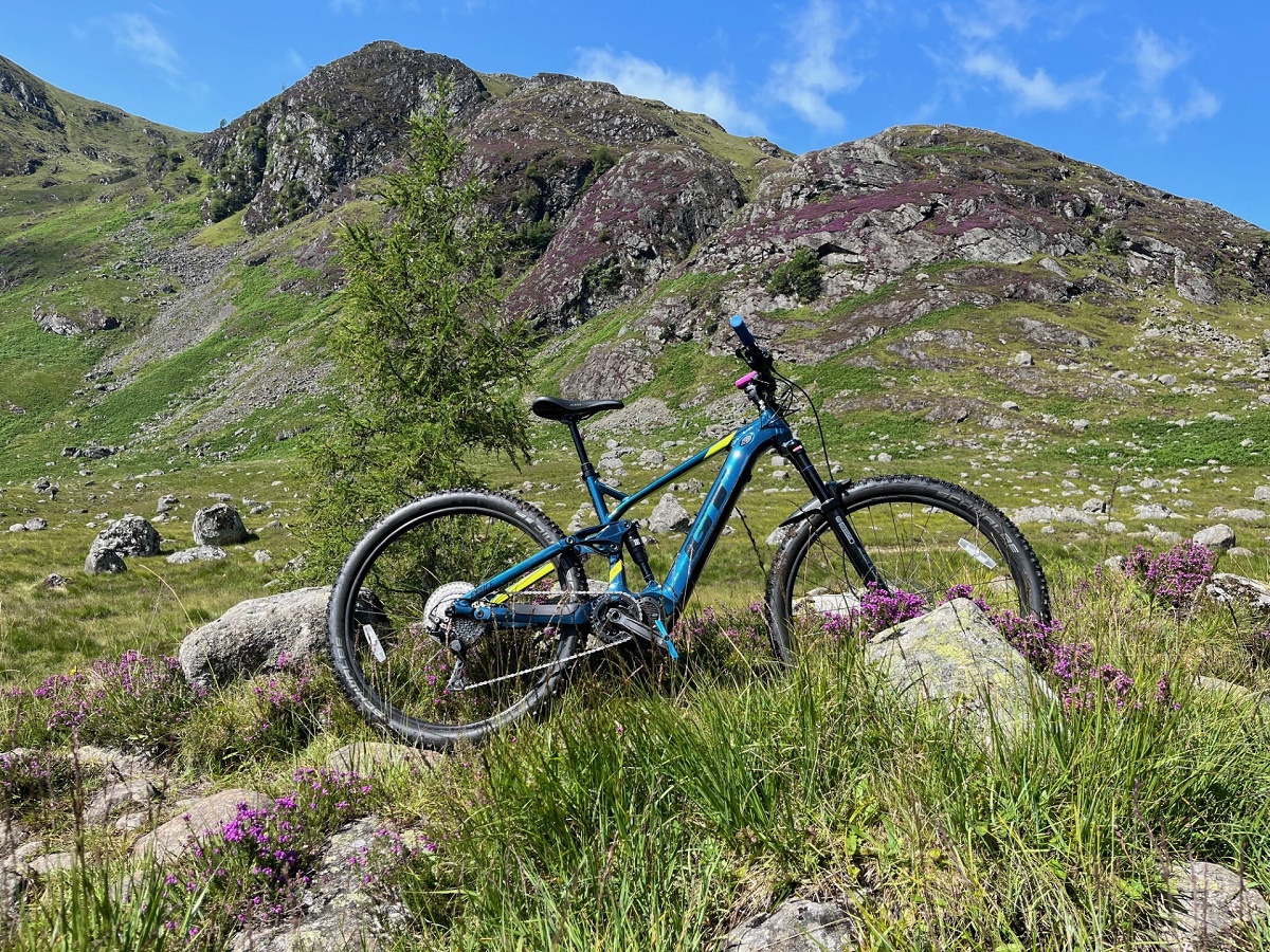 A blue and yellow mountain bike placed among rocks and purple heather in a rugged mountainous landscape.