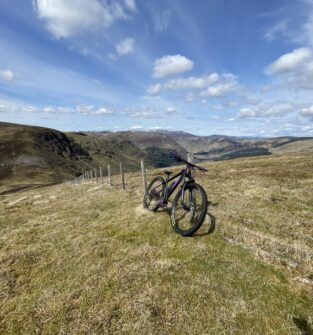 A purple mountain bike resting against a wire fence on a grassy hillside, overlooking a broad valley under a blue sky with white clouds.